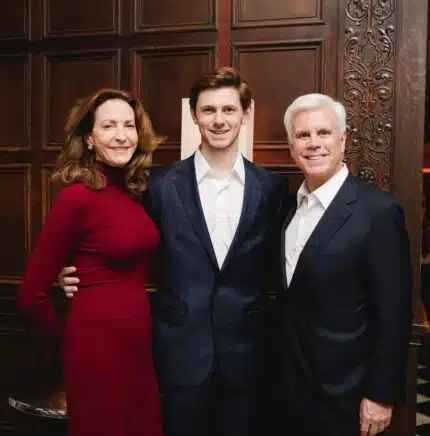 Alex Norcross stands with his parent on either side of him. All are wearing professional clothing and smiling, looking at camera.