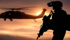 An armed soldier in silhouette with a helicopter hovering on his left.