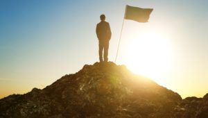 Man at the summit of a mountain with the sun behind him and a flag planted in the ground.