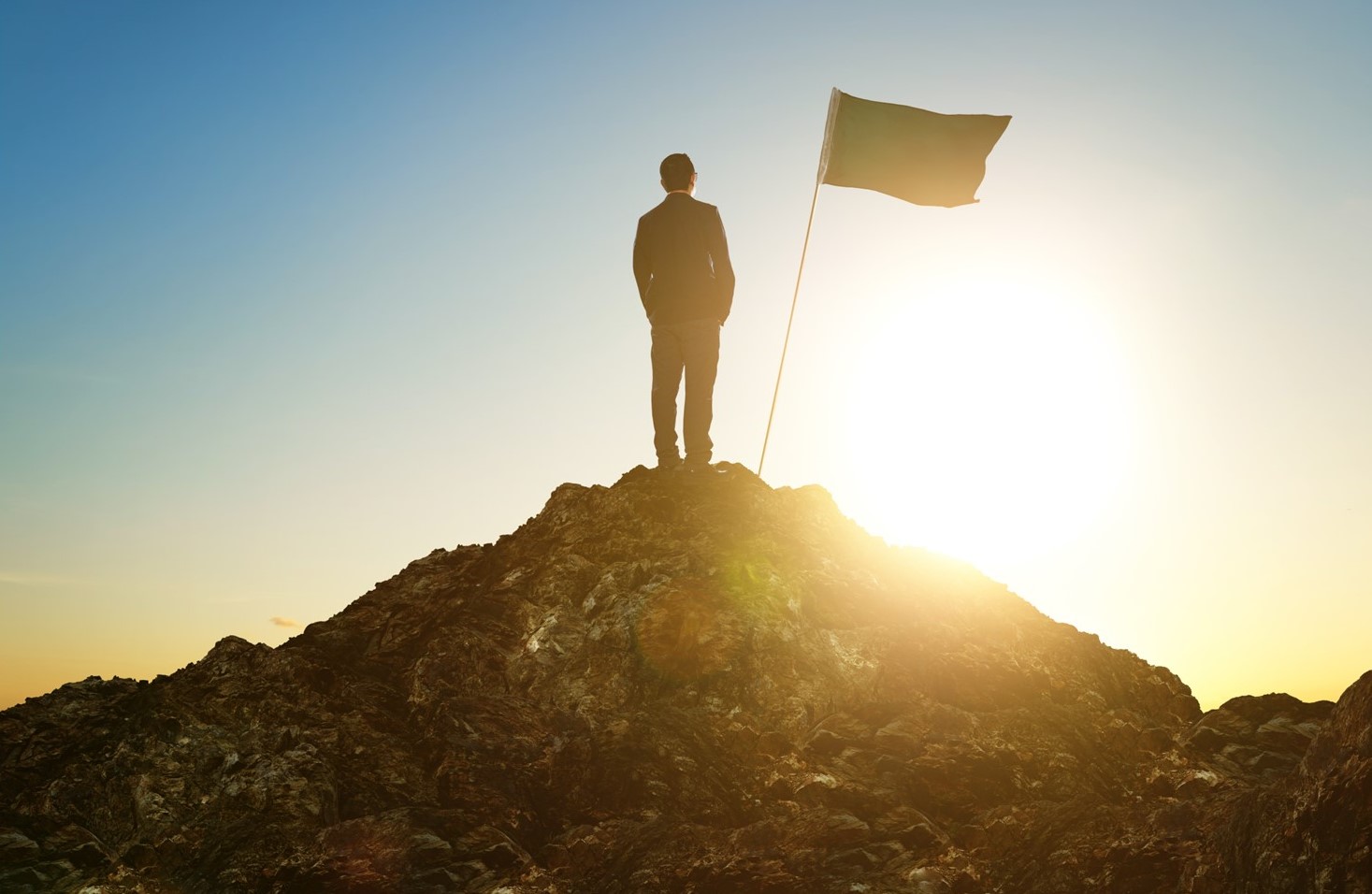 Man at the summit of a mountain with the sun behind him and a flag planted in the ground.