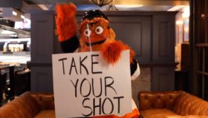 Gritty, holding a "Take Your Shot" sign, will be visiting Radnor High for a Flyers vaccine promotion.