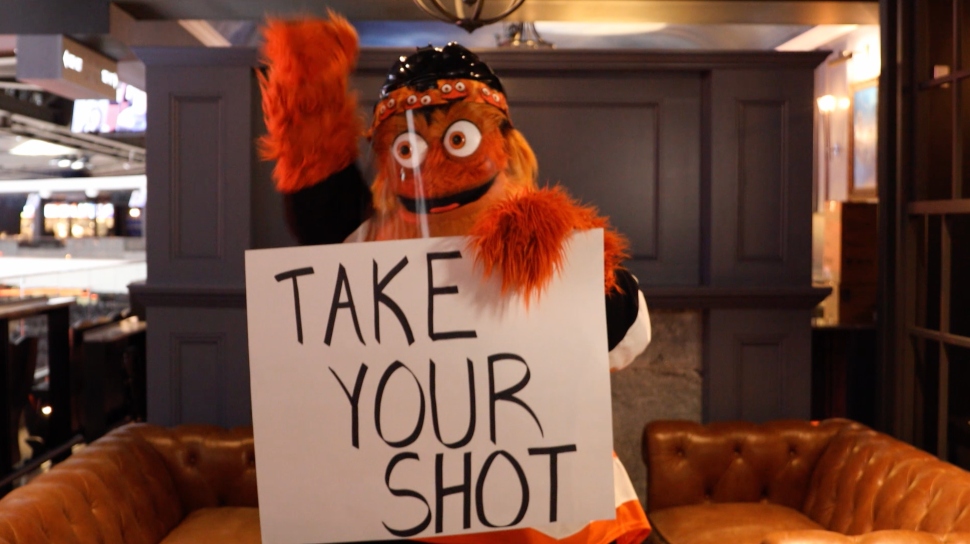 Gritty, holding a "Take Your Shot" sign, will be visiting Radnor High for a Flyers vaccine promotion.