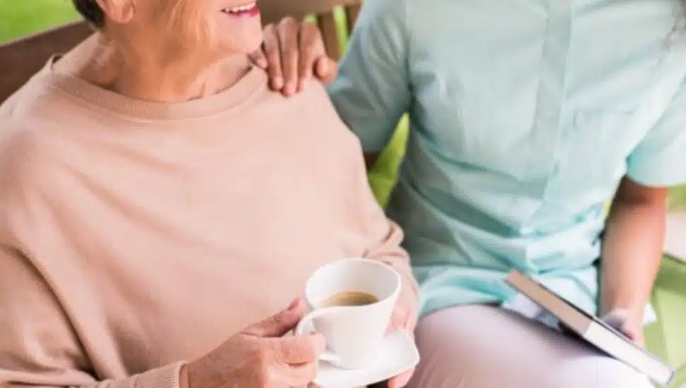 A senior citizen sitting with a cup of coffee talking to another person next to them.