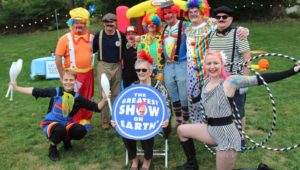 Woman holds "The Greatest Show on Earth" sign, surrounded by peoople dressed as circus performers.