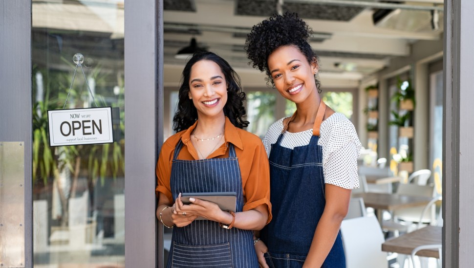 Happy successful multiethnic small business women wearing apron and standing with open sign at entrance gate of restaurant.