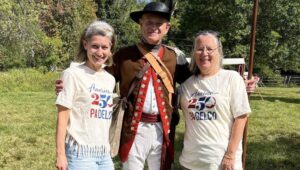 Two women with America250PADelco T-shirts standing with a Revolutionary War soldier