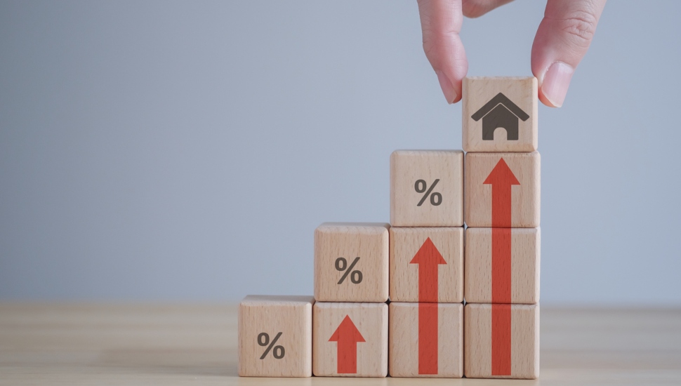 Hand holding house icon on wooden block from stack of block with percent and rise of arrow.