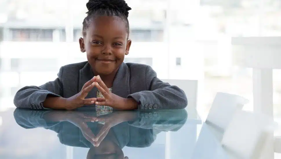A smiling young Black child with hand clasped sitting at a table