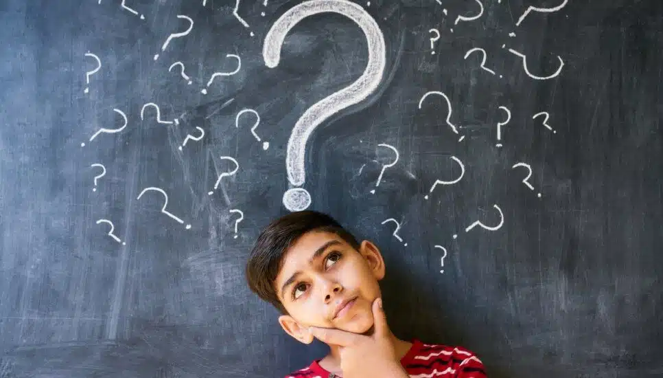 An uncertain boy student at the blackboard surrounded by question marks