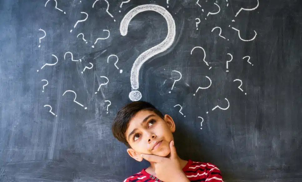 An uncertain boy student at the blackboard surrounded by question marks