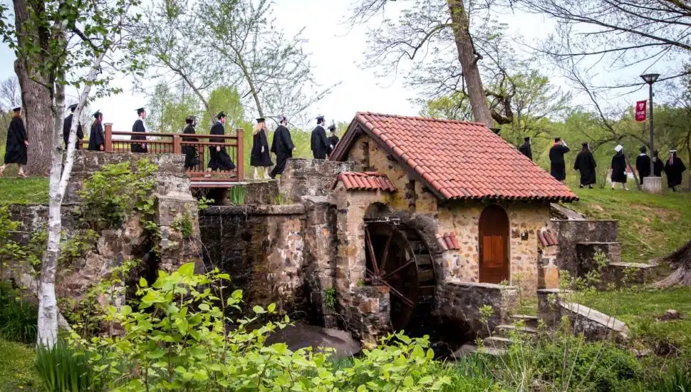 Seniors in cap and gown on the main campus of Eastern University walking past a water mill