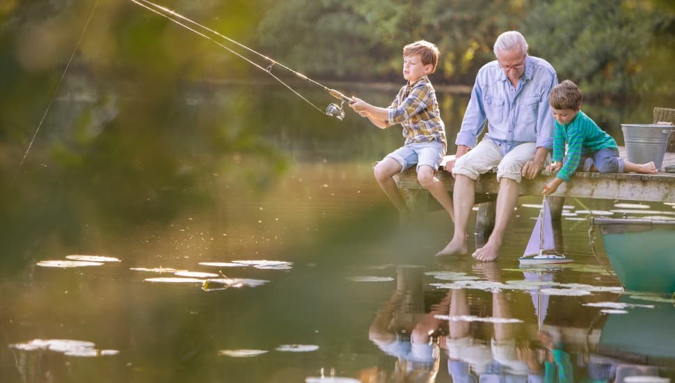 An older man is fishing by a creek or river with two young boys.