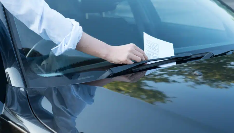 A hand reaches for a parking ticket placed on the windshield of a car