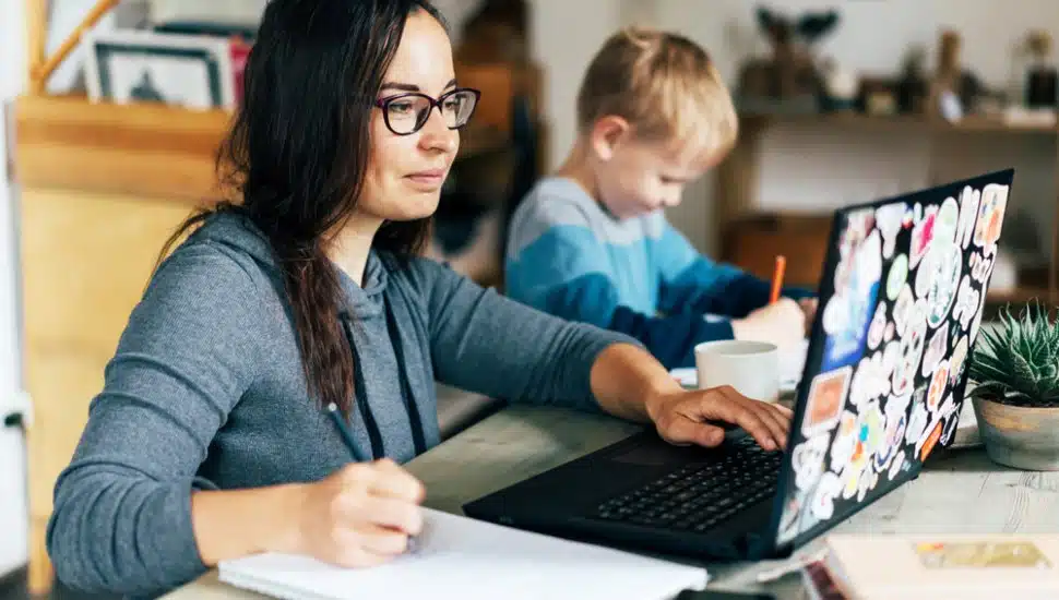 A woman looking at a laptop computer while writing on a tablet, with her young son nearby.