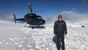 Jere Longman stands alone on top of a snowy mountain with a back helicopter behind him.