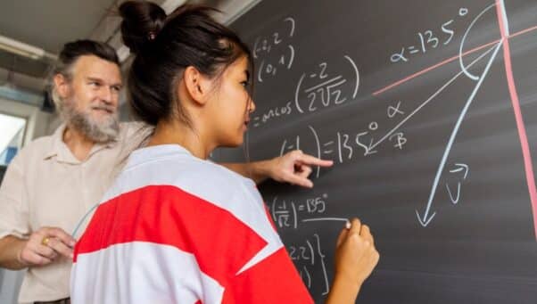 A student works with a teacher on a math problem shown on a black board.