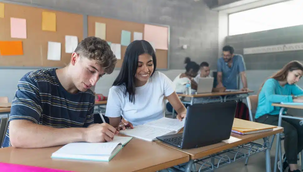 Two students, a boy and a girl, are sharing study time in a classroom taking notes and looking at a computer screen.