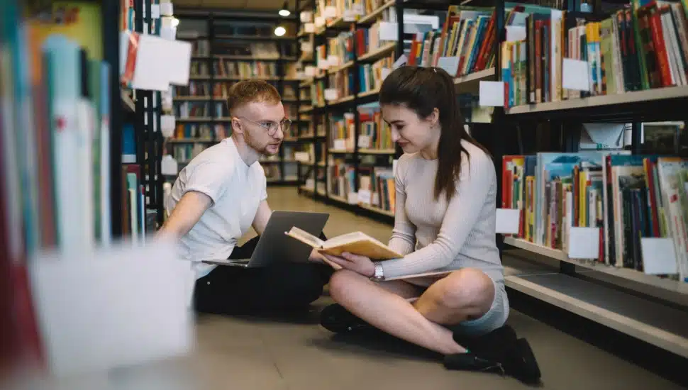 Two college students look through a book while sitting on the floor of a library.