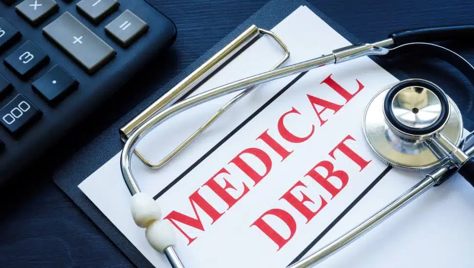 A clip board with a medical debt bill, a stethoscope and a calculator.
