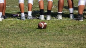 A football rests on the field as a football team lines up near it.