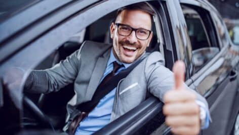 A man in a suit behind the wheel of a car giving the thumbs up out of his driver's side window.