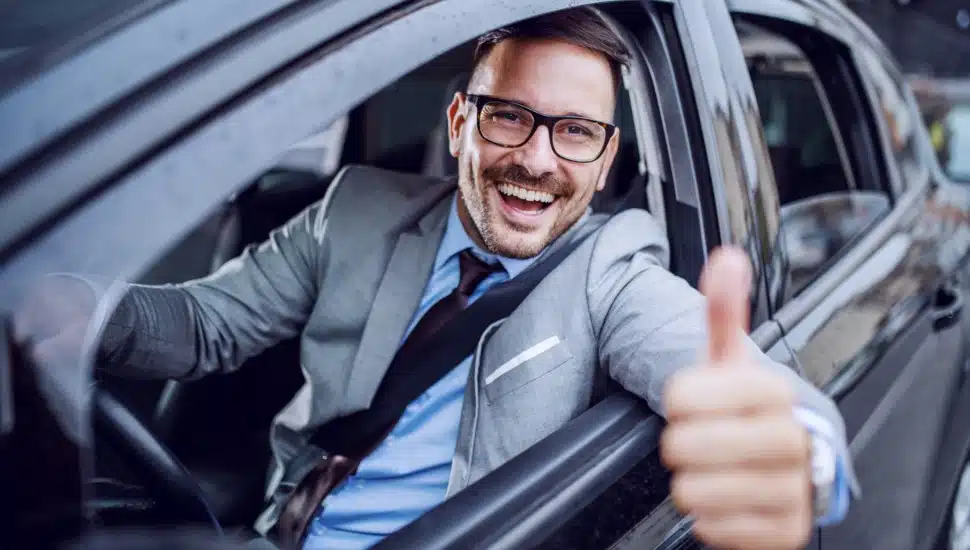 A man in a suit behind the wheel of a car giving the thumbs up out of his driver's side window.