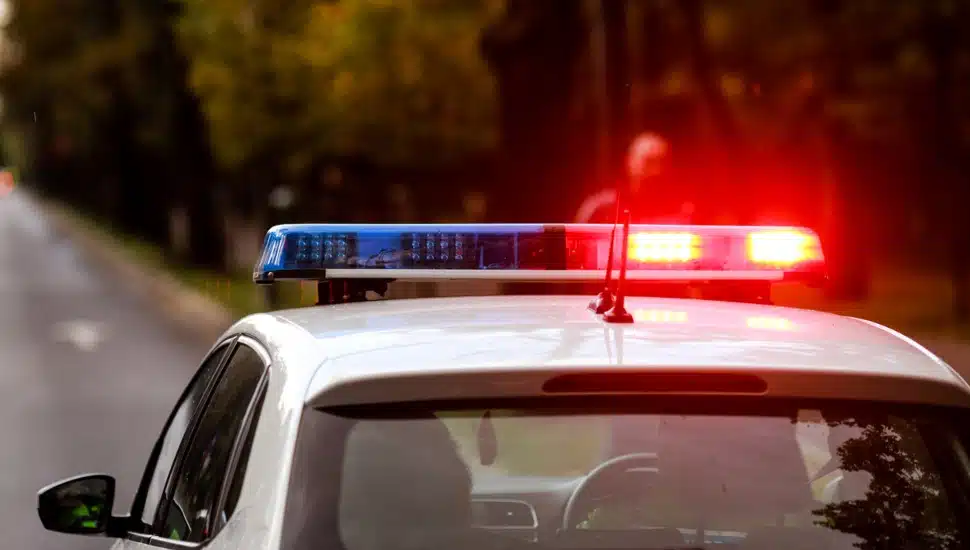 A police car with its lights running on an open road approaching a man walking.