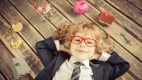 A child in a business suit relaxes, surrounded by a piggy bank, scales, and model homes.