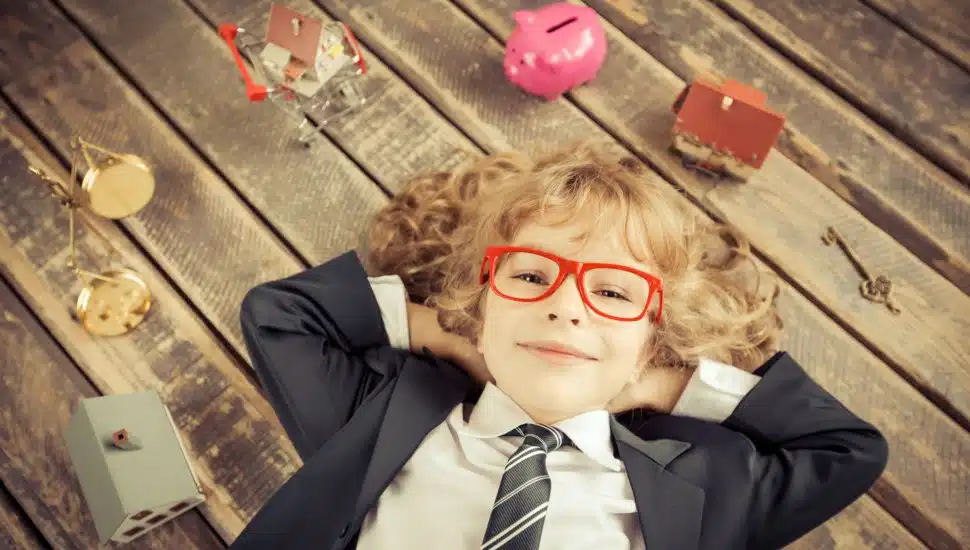 A child in a business suit relaxes, surrounded by a piggy bank, scales, and model homes.