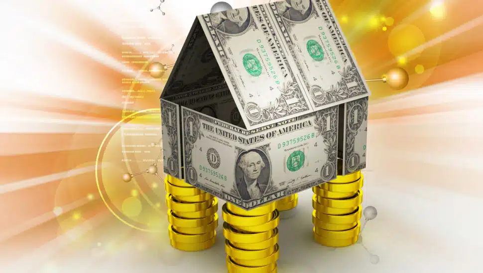 A miniature home made out of dollar bills, propped up by gold coins.