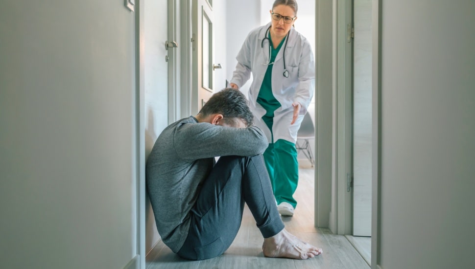 A worried female psychiatrist runs to help a male patient with mental disorder sitting on the floor.