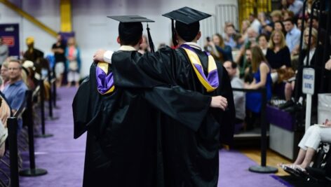a graduation ceremony at WCU with two graduates with in cap and gown with arms around each other's shoulders.