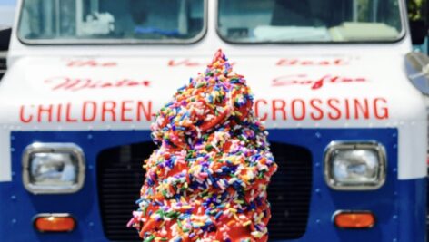 An ice cream cone with sprinkles in the foreground, with the front of an ice cream truck in the background.