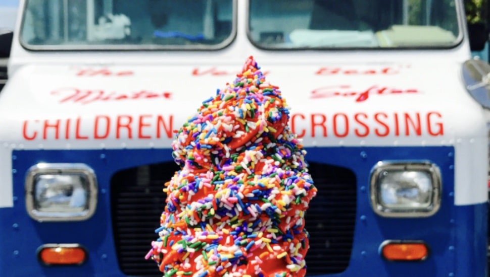 An ice cream cone with sprinkles in the foreground, with the front of an ice cream truck in the background.