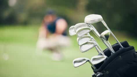 A bag of golf clubs in the foreground with a golfer prepping to play blurred out in the distance.