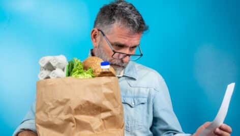 A man holds a bag of groceries while looking at a bill.