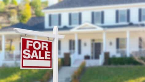 A for sale sits on the lawn of a large home in the suburbs.