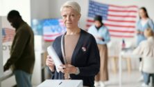 Mature blond female citizen of USA looking at camera while standing by box and holding ballot paper with her choice of candidate.