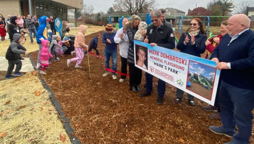 Officials hold a sign as kids play at a ribbon cutting for the Mark Dombroski Memorial Playground in Middletown.