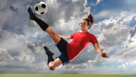 A woman soccer player kicks jumps in the air as she connects her foot with the ball.