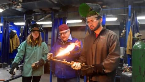 A trainer demonstrates a welding technique in front of two students.