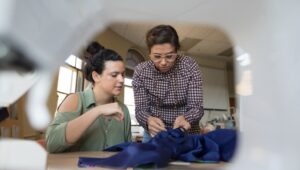 Two women collaborating on fabric design in a creative workspace, viewed through the opening of a sewing machine.