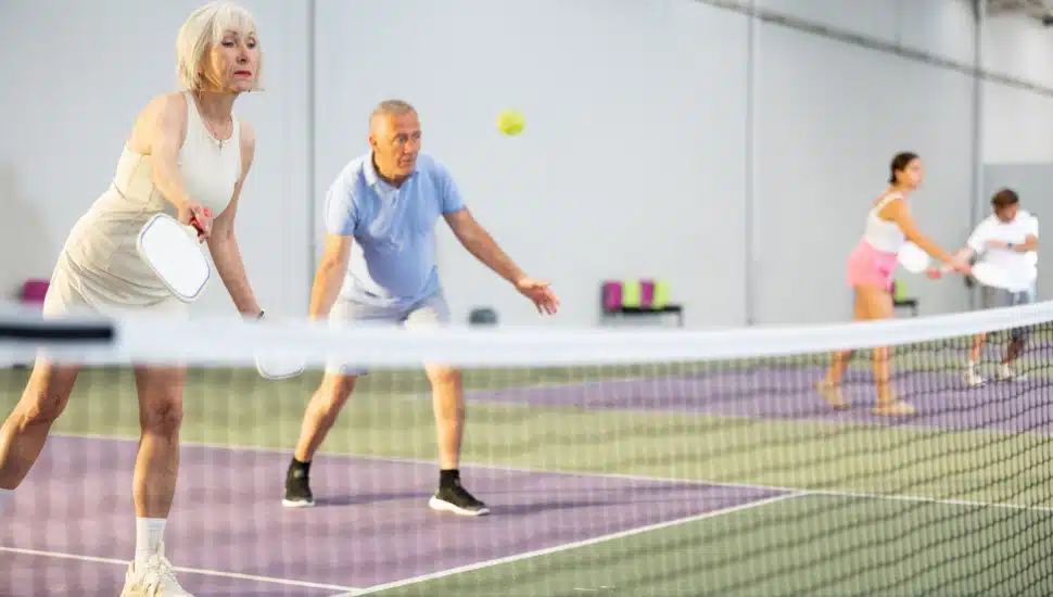 An elderly woman plays pickleball with a male partner on an indoor court.