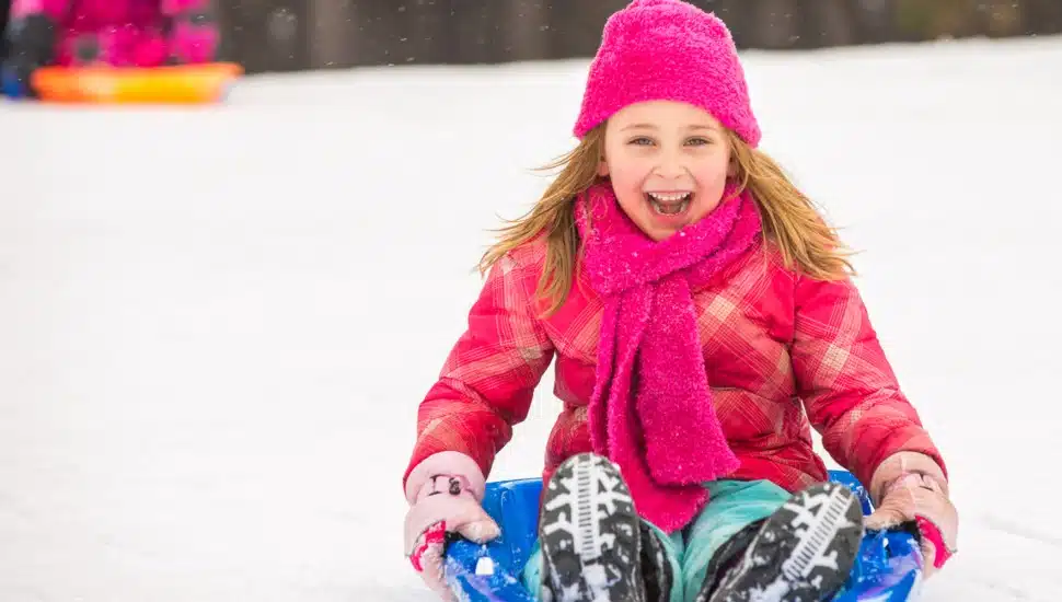 A girl with pink clothing sleds down a snow-covered hill.
