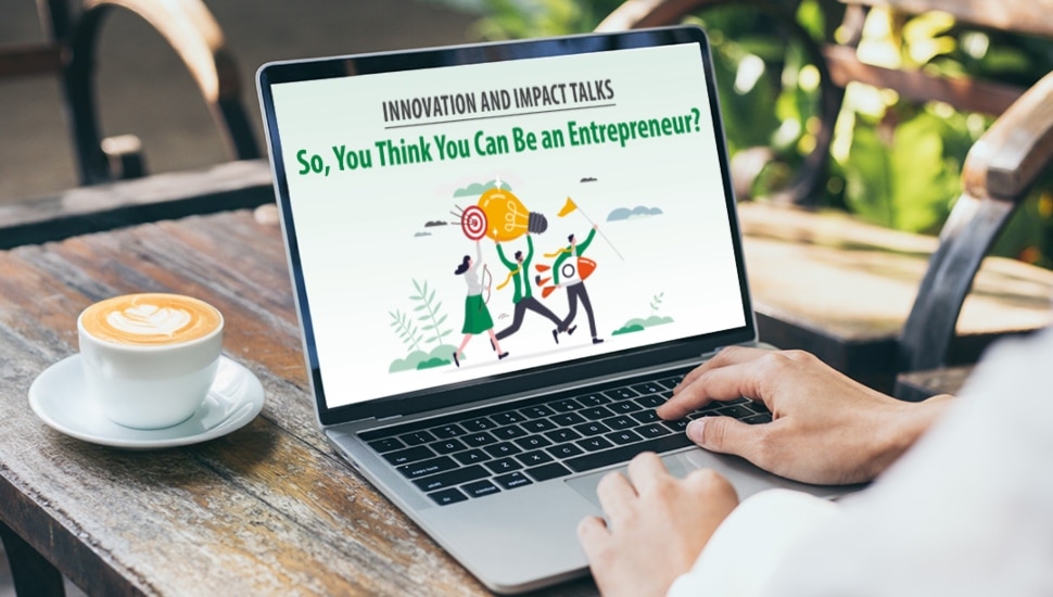 A person using a laptop at an outdoor café with a cup of coffee beside them. The laptop screen displays a promotional graphic for Wilmington University’s Innovation and Impact Talks event titled "So, You Think You Can Be an Entrepreneur?" featuring an illustration of people carrying a giant lightbulb, symbolizing innovation and entrepreneurship.