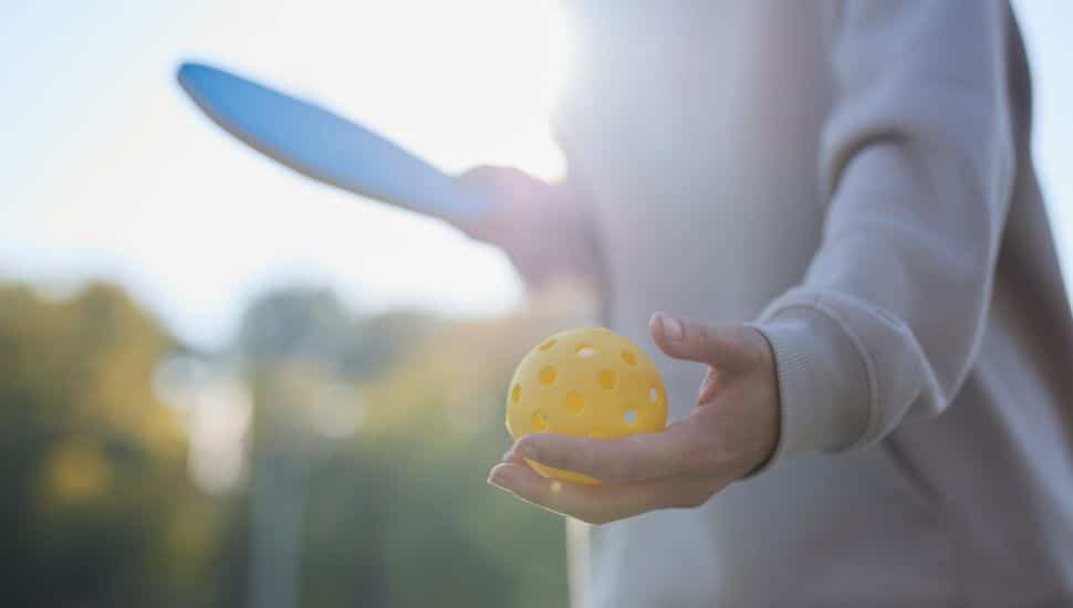 A person with a pickleball and paddle gets ready to serve.