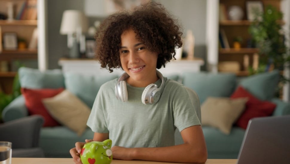 A smiling teenage girl wearing headphones holds a piggy bank.