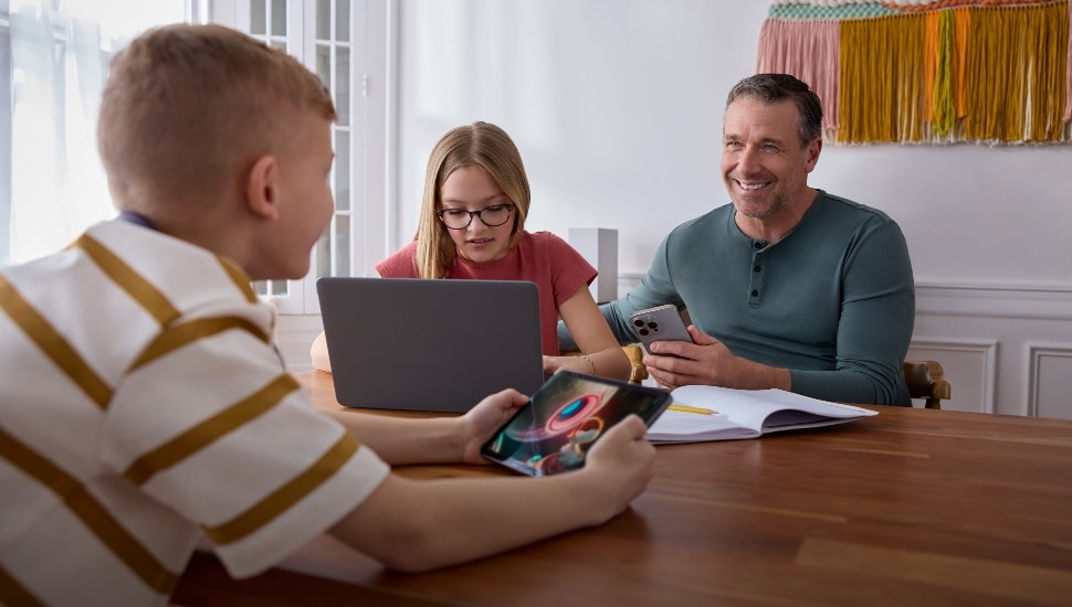 Three people sit at a table. A woman is on her laptop. The two men are using mobile devices.