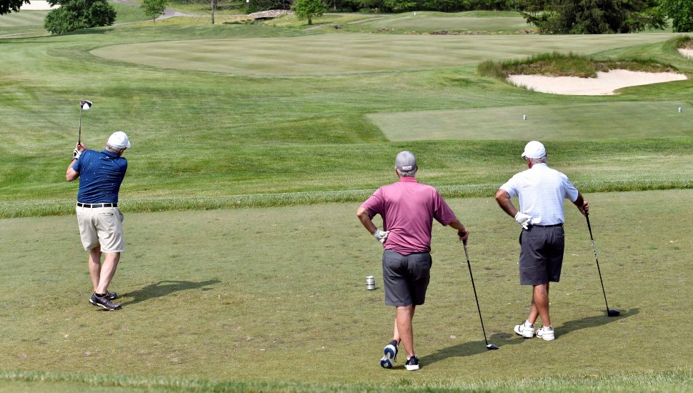 Three golfers pictured at a distance playing a game of golf. One is swinging, the other two are watching.
