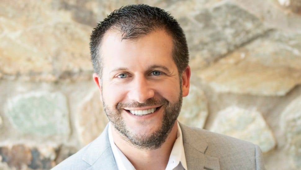 Headshot of Dave Batty in front of a stone wall. Batty is wearing a gray blazer and white collared shirt. He has brown hair and a brown beard and is smiling.